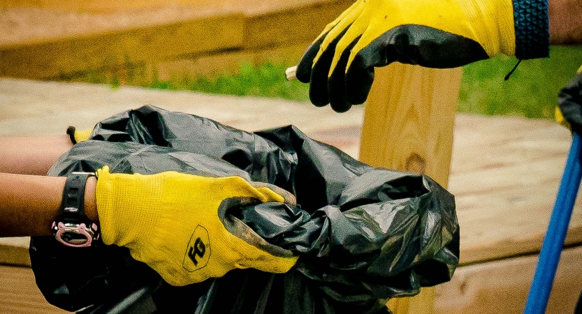 Gloved hands hold open a black trash bag while another gloved hand drops something into the bag.